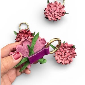 Op de afbeelding: Roze en bordeauxrode leren bloemvormige sleutelhangers met goudkleurige ringen en groene bladeren. E&eacute;n sleutelhanger wordt door een hand vastgehouden. De sleutelhangers staan op een witte achtergrond.