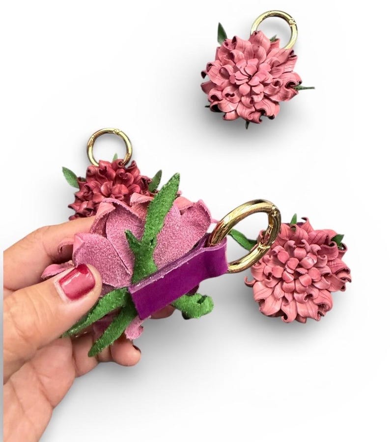 Op de afbeelding: Roze en bordeauxrode leren bloemvormige sleutelhangers met goudkleurige ringen en groene bladeren. E&eacute;n sleutelhanger wordt door een hand vastgehouden. De sleutelhangers staan op een witte achtergrond.