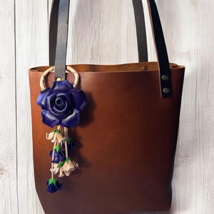 May include: A brown leather tote bag with black leather straps and a blue leather rose charm hanging from the handle.