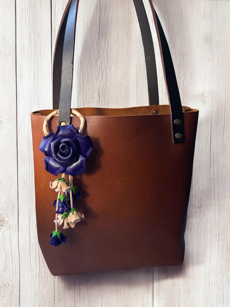 May include: A brown leather tote bag with black leather straps and a blue leather rose charm hanging from the handle.