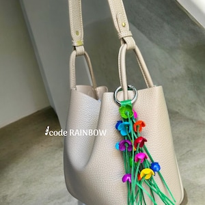 K&ouml;nnte beinhalten: Eine beige Ledertasche mit einem langen Riemen und einem bunten Blumenanh&auml;nger. Der Anh&auml;nger hat gr&uuml;ne Stiele und mehrfarbige Blumen.