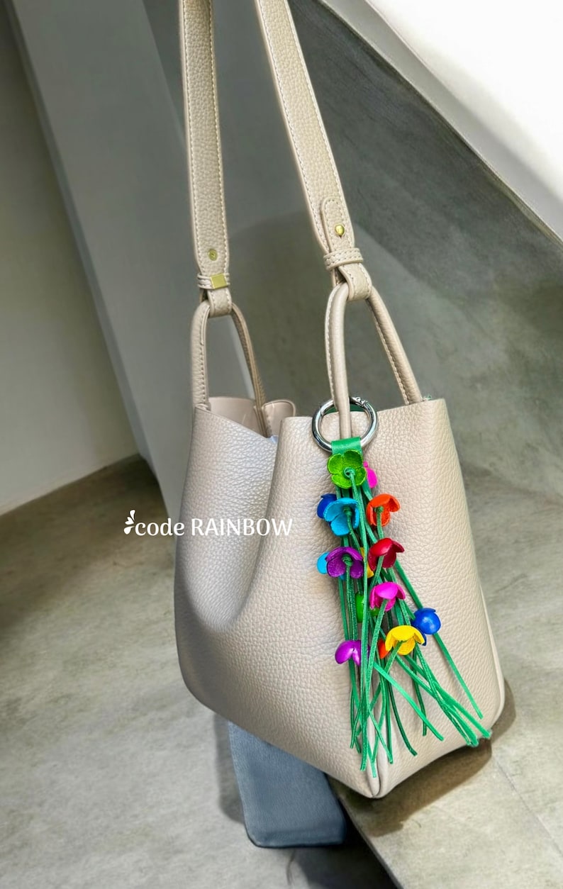 K&ouml;nnte beinhalten: Eine beige Ledertasche mit einem langen Riemen und einem bunten Blumenanh&auml;nger. Der Anh&auml;nger hat gr&uuml;ne Stiele und mehrfarbige Blumen.
