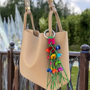 Peut inclure: Un sac cabas en cuir beige avec un charme de fleur coloré. Le charme a des tiges vertes et des fleurs multicolores.