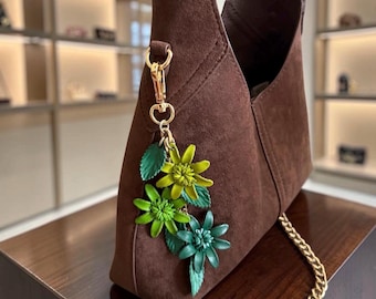 Könnte beinhalten: Eine dunkelbraune Wildleder-Hobo-Tasche mit einem goldfarbenen Metallverschluss, an dem ein dekorativer Anhänger befestigt ist. Der Anhänger besteht aus drei geschichteten, grün-gelben Blüten mit grünen Blättern und einem goldfarbenen Kettenriemen.