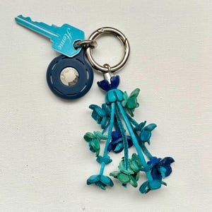 May include: A blue and green leather flower keychain with a silver ring and a blue round charm with the word "Jeanne" on a silver key.