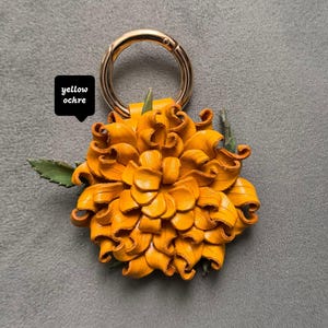 Op de afbeelding: Een sleutelhanger met een gele okerkleurige leren bloem met een goudkleurige metalen ring. De bloem heeft een gedetailleerde bloembladconstructie en groene bladaccenten. De tekst "yellow ochre" staat in een zwart vierkant.