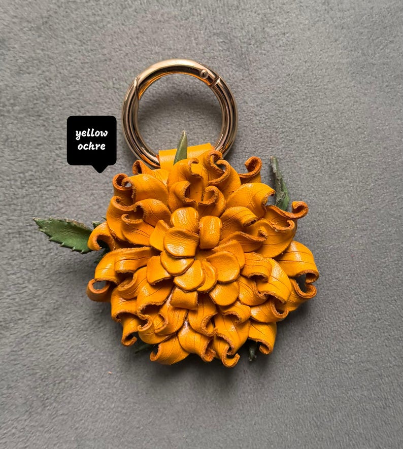 Op de afbeelding: Een sleutelhanger met een gele okerkleurige leren bloem met een goudkleurige metalen ring. De bloem heeft een gedetailleerde bloembladconstructie en groene bladaccenten. De tekst "yellow ochre" staat in een zwart vierkant.