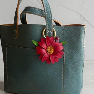 May include: Teal leather tote bag with a red flower accent. The bag has a small pocket on the front and gold-colored rings connecting the teal handles. The flower has a yellow center and a small ladybug.