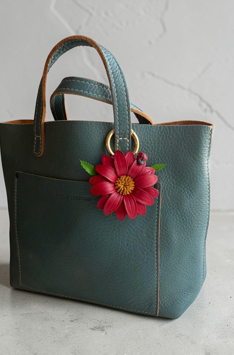 May include: Teal leather tote bag with a red flower accent. The bag has a small pocket on the front and gold-colored rings connecting the teal handles. The flower has a yellow center and a small ladybug.