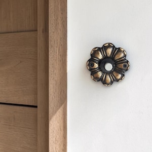 May include: A decorative, antique-style doorbell button with a floral design. The button is made of bronze-colored metal and is mounted on a white wall. The doorbell button is near a wooden door frame.