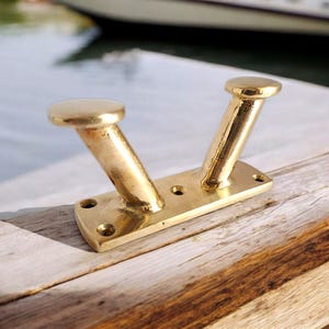 May include: A polished brass boat cleat featuring two upward-angled horns and a rectangular base plate with four pre-drilled screw holes. The metallic surface reflects light, showing a smooth, shiny finish on the hardware mounted to wood.