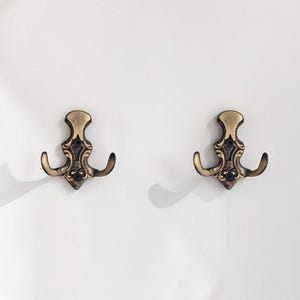 May include: Two antique-style brass wall hooks with a decorative design. Each hook has a double prong and a detailed top, perfect for hanging coats, hats, or other items. The hooks are mounted on a white wall, showcasing their ornate details.
