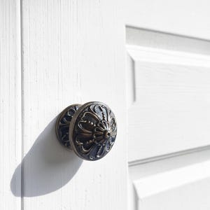 May include: A round, dark bronze-toned metal cabinet knob featuring an intricate, raised floral-inspired relief pattern with a central point and curved petals, mounted on a white paneled door with a visible vertical wood grain texture.