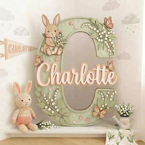 May include: A light green letter "C" decoration featuring a bunny, butterflies, and the name "Charlotte". The design includes floral elements and is complemented by a stuffed bunny and a pennant, perfect for a nursery.