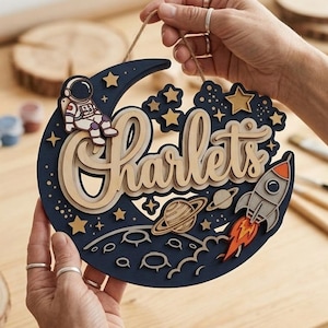 Può includere: Un'insegna in legno a strati a tema spaziale in blu navy, beige e oro. L'insegna presenta una luna crescente, stelle, pianeti e un razzo. Il nome "Charlets" è scritto in corsivo.