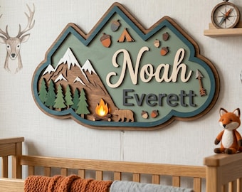 May include: A wooden nursery name sign in a mountain theme. The sign features the names "Noah" and "Everett" in a layered design with mountains, trees, a campfire, and a bear. The color palette includes green, brown, and white.