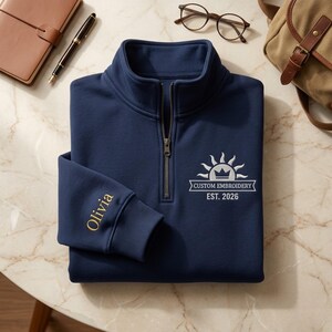 May include: A navy blue quarter-zip sweatshirt featuring white embroidery of a sun icon with a crown and the text "CUSTOM EMBROIDERY EST. 2026" on the chest. The sleeve is embroidered with the name "Olivia" in gold thread.