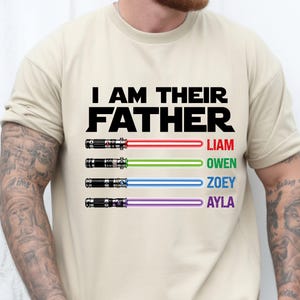 Pu&ograve; includere: T-shirt beige con la scritta nera "I AM THEIR FATHER". Sotto, quattro spade laser con i nomi Liam, Owen, Zoey e Ayla in rosso, verde, blu e viola.