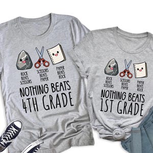 May include: Two heather gray t-shirts featuring a rock, scissors, and paper graphic with the text "Rock beats scissors, scissors beats paper, paper beats rock" and "Nothing beats 4th grade" or "Nothing beats 1st grade" alongside two black sneakers and blue jeans.
