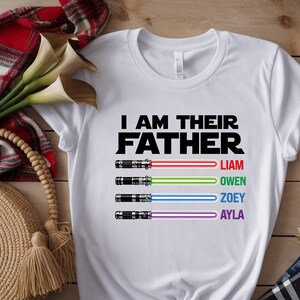 Pu&ograve; includere: Maglietta bianca con la scritta nera "I AM THEIR FATHER". Sotto, quattro spade laser con raggi rossi, verdi, blu e viola, ciascuna con un nome: Liam, Owen, Zoey e Ayla.