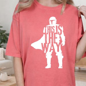 以下が含まれることがあります： コーラルカラーの半袖Tシャツ。ヘルメットを被り鎧を着たキャラクターの白いシルエットが描かれています。グラフィックには「THIS IS THE WAY」という文字がキャラクターの体の一部として組み込まれており、かすれたようなヴィンテージ風の質感が特徴です。