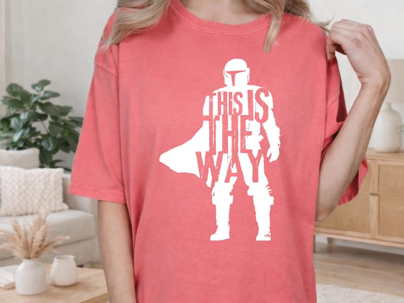 以下が含まれることがあります： コーラルカラーの半袖Tシャツ。ヘルメットを被り鎧を着たキャラクターの白いシルエットが描かれています。グラフィックには「THIS IS THE WAY」という文字がキャラクターの体の一部として組み込まれており、かすれたようなヴィンテージ風の質感が特徴です。
