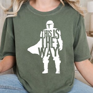 以下が含まれることがあります： オリーブグリーンの半袖Tシャツ。ヘルメットを被り鎧を着てマントを羽織ったキャラクターの白いシルエットが描かれています。キャラクターの胴体中央には「THIS IS THE WAY」という文字が大きな白い大文字でプリントされています。