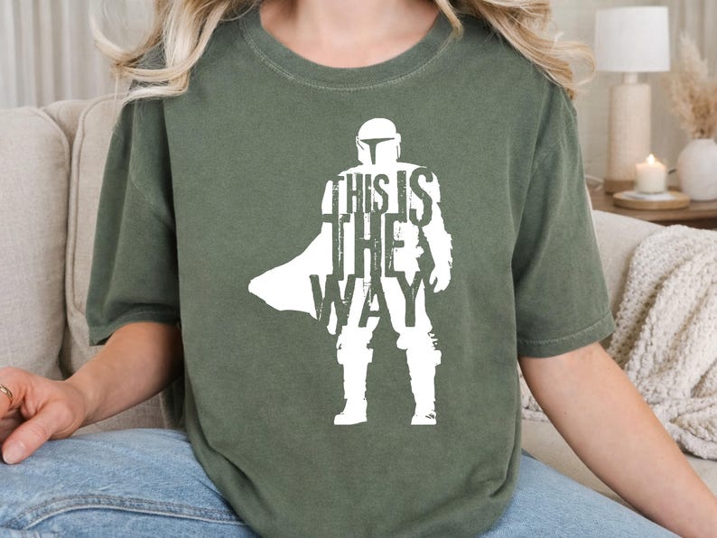 以下が含まれることがあります： オリーブグリーンの半袖Tシャツ。ヘルメットを被り鎧を着てマントを羽織ったキャラクターの白いシルエットが描かれています。キャラクターの胴体中央には「THIS IS THE WAY」という文字が大きな白い大文字でプリントされています。
