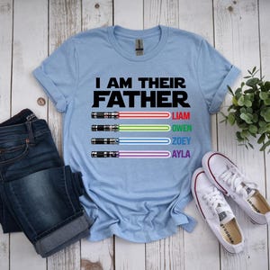 Pu&ograve; includere: Maglietta azzurra con la scritta "I AM THEIR FATHER" e disegni di spade laser con nomi. L'immagine comprende jeans blu e scarpe da ginnastica bianche. &Egrave; visibile anche una piccola pianta verde.