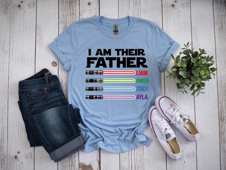 Pu&ograve; includere: Maglietta azzurra con la scritta "I AM THEIR FATHER" e disegni di spade laser con nomi. L'immagine comprende jeans blu e scarpe da ginnastica bianche. &Egrave; visibile anche una piccola pianta verde.