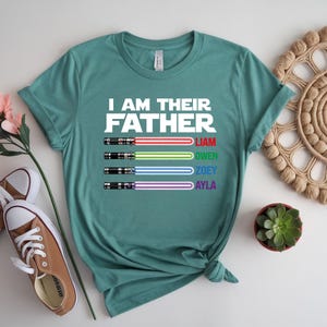 Pu&ograve; includere: T-shirt verde acqua con la scritta "I AM THEIR FATHER" e quattro disegni di spade laser colorate. Sotto, i nomi Liam, Owen, Zoey e Ayla. La maglietta &egrave; presentata con scarpe da ginnastica marroni e un fiore.