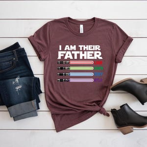 Pu&ograve; includere: Una maglietta bordeaux con la scritta "I AM THEIR FATHER" e quattro disegni di spade laser rosse, verdi, blu e viola. I nomi Liam, Owen, Joey e Ayla sono sotto. Jeans scuri e stivaletti neri sono anche nell'immagine.