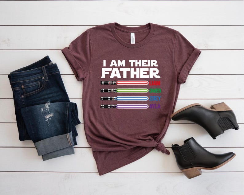 Pu&ograve; includere: Una maglietta bordeaux con la scritta "I AM THEIR FATHER" e quattro disegni di spade laser rosse, verdi, blu e viola. I nomi Liam, Owen, Joey e Ayla sono sotto. Jeans scuri e stivaletti neri sono anche nell'immagine.
