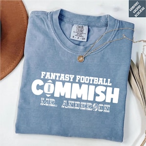 May include: A blue short-sleeved t-shirt with a white graphic print that reads FANTASY FOOTBALL COMMISH and MR. ANDERSON, featuring a football icon in place of the letter O. The shirt has a crew neckline and a Comfort Colors brand label inside.