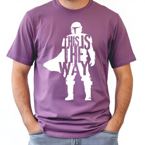 以下が含まれることがあります： 紫色の半袖Tシャツで、ヘルメットを被り鎧を着てマントを羽織ったキャラクターの白いグラフィックプリントが施されています。キャラクターのシルエットの中央には、「THIS IS THE WAY」というテキストがかすれたフォントスタイルで白くプリントされています。