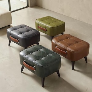 May include: Four tufted ottomans in various colors: dark gray, olive green, brown, and deep teal. Each has a rectangular shape, wooden legs, and a brown leather handle. The ottomans are arranged on a light-colored floor.