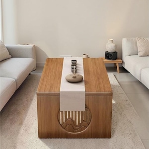 May include: A rectangular wooden coffee table with a light brown finish. The table has a decorative carved design on the side and a beige runner with tea set. The table is placed on a light-colored rug in a living room setting.