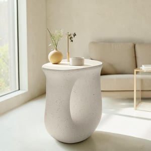 Puede incluir: Una mesa auxiliar moderna, de color blanco roto, con un diseño curvo único. La mesa tiene una parte superior circular y una abertura distintiva en el lateral. Un pequeño jarrón redondo, una pieza decorativa dorada y un cuenco blanco pequeño están encima. La mesa mide unos 61 cm de alto.