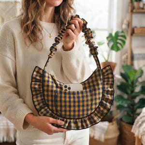 Puede incluir: Un bolso de mano en forma de media luna con un patrón de cuadros vichy en amarillo y azul marino. El bolso tiene un borde con volantes y una correa de hombro fruncida estilo coletero. Un smartphone blanco está en el bolsillo exterior.
