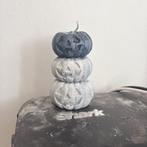 Puede incluir: Una pila de tres velas con forma de calabaza tallada en un degradado de colores, que va desde el gris carbón oscuro en la parte superior hasta un gris pálido más claro en la base. Cada calabaza presenta una cara de linterna con ojos triangulares.