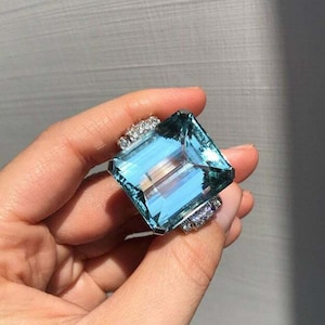 Può includere: Un anello con una grande pietra di acquamarina a taglio quadrato. La pietra blu chiaro è incastonata in una fascia d'argento con pietre più piccole ai lati. L'anello è tenuto su uno sfondo grigio.
