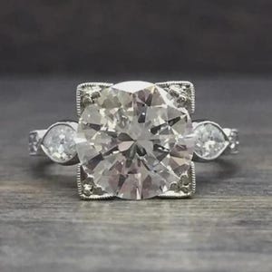 May include: A silver engagement ring with a large, round, clear gemstone at the center, flanked by two smaller pear-shaped stones. The center stone is set in a square frame with decorative detailing.