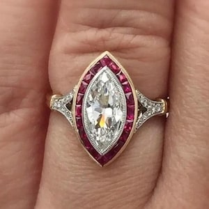May include: A marquise-cut diamond ring set in yellow gold, featuring a halo of vibrant red rubies surrounding the center stone. The band includes intricate metalwork with small accent diamonds along the shoulders of the ring setting.
