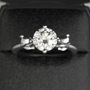 May include: A silver engagement ring with a large, round, clear gemstone as the centerpiece. The ring is set with two smaller gemstones on either side of the main stone. The ring is displayed in a black ring box.