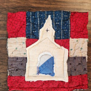 May include: A square quilted fabric patch featuring a central white fabric applique of a church building with a small cross. The background is composed of a patchwork grid of red, dark blue, and off-white patterned fabric squares with visible stitching.