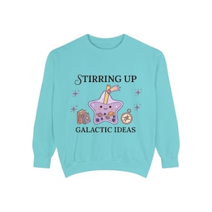 Puede incluir: Una sudadera de manga larga azul claro con el texto "Stirring Up Galactic Ideas". El gráfico presenta una bebida boba en forma de estrella, libros, una brújula y estrellas. El diseño es en tonos morados, amarillos y marrones.