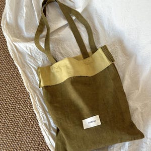 Op de afbeelding: Olijfgroene draagtas met een contrasterende, beige bovenrand en bijpassende handvatten. Een klein wit label met de tekst "SolBlur" is op de voorkant genaaid. De tas is gemaakt van een stevige stof.