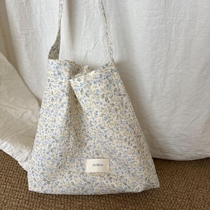 May include: A tote bag made of white fabric with a small blue and green floral pattern. The bag features a single shoulder strap and a rectangular white fabric label on the front with the black text SolBlur printed in the center.