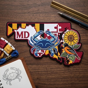 May include: Embroidered patch in the shape of Maryland, featuring a blue crab, sunflower, and Baltimore oriole. The patch includes the letters "MD" and is bordered in black and red. A sketch of a crab is in a notebook.