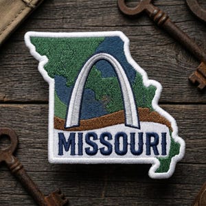 May include: Embroidered patch in the shape of Missouri, featuring the Gateway Arch and the word "MISSOURI" in navy blue. The patch has a white border and is set against a wooden background with antique keys.